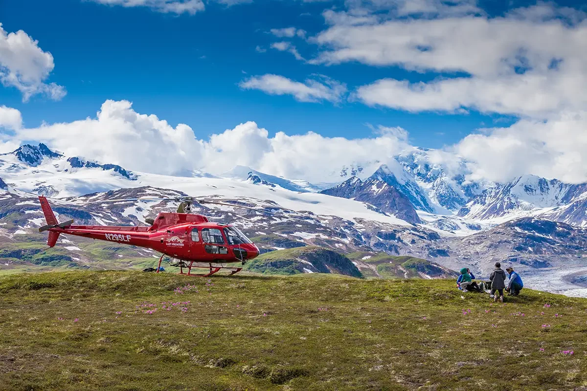 Three people hitchiking in the Alaskan mountains next to a red helicopter